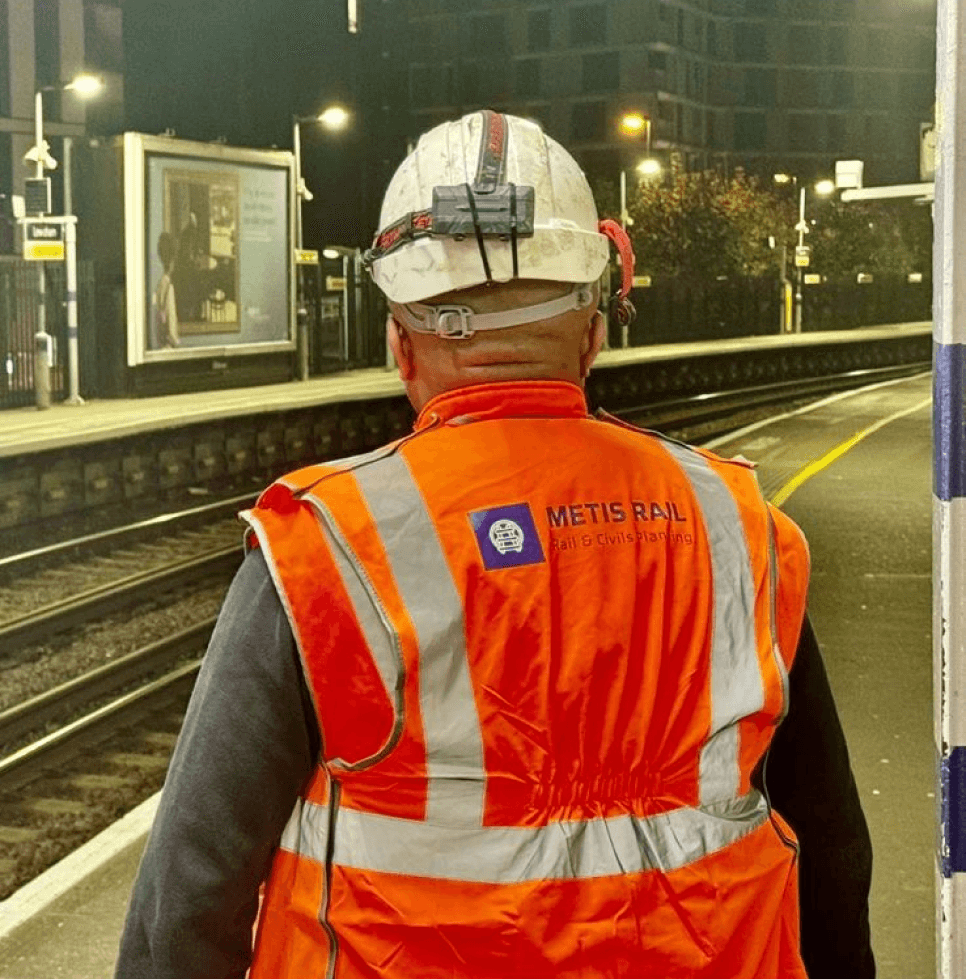 Man wearing orange Metis Rail high-visibility safety gear and white hard hat standing on a train platform, with his back to the camera