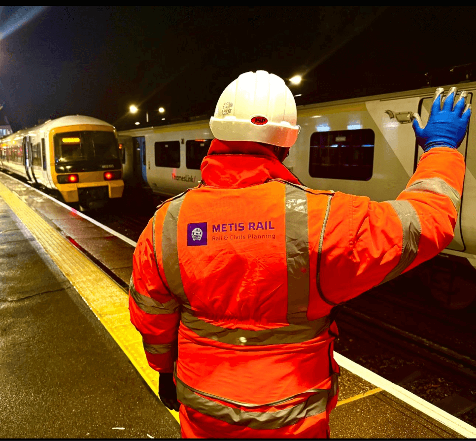 A man on a train station platform, wearing a bright orange Metis Rail uniform, hand signalling to a train pulling into the station.
