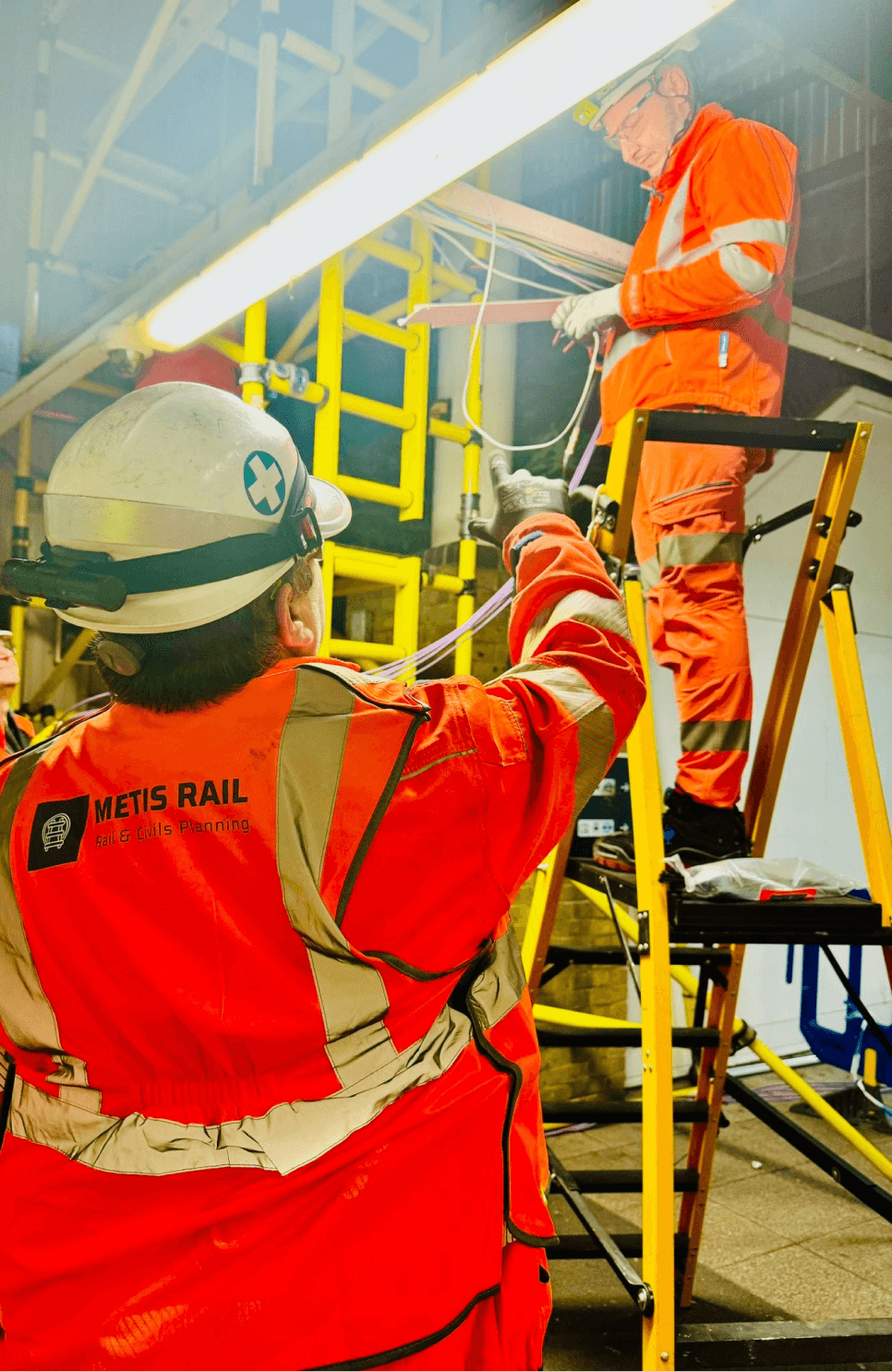 Two Metis Rail workers in high-visibility safety gear performing electrical work on-site, with one on a ladder and the other providing guidance.